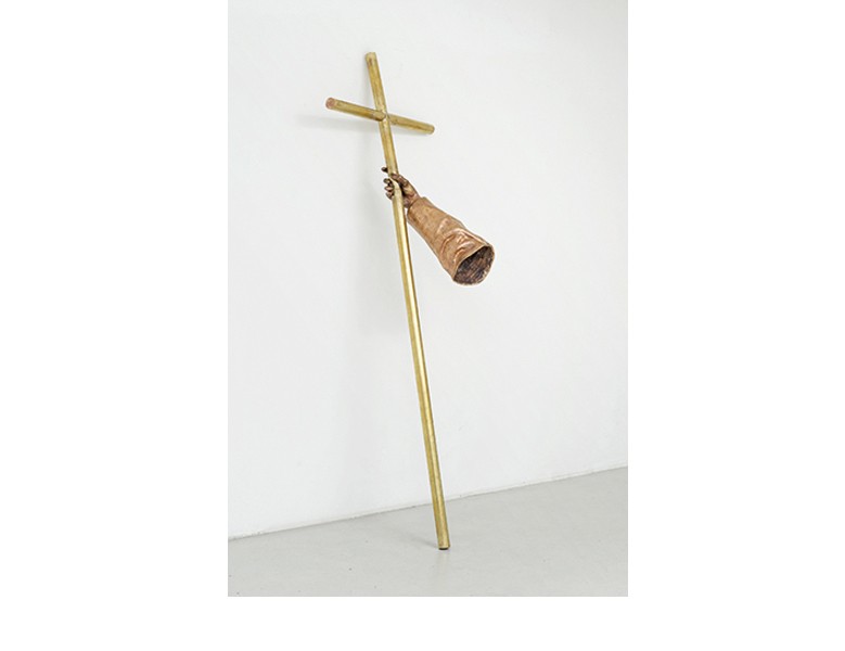 Sculpture by Fayçal Baghriche titled "Le bras du Cardinal (The Cardinal's Arm)." A bronze for arm holds a brass cross which leans against a white wall.