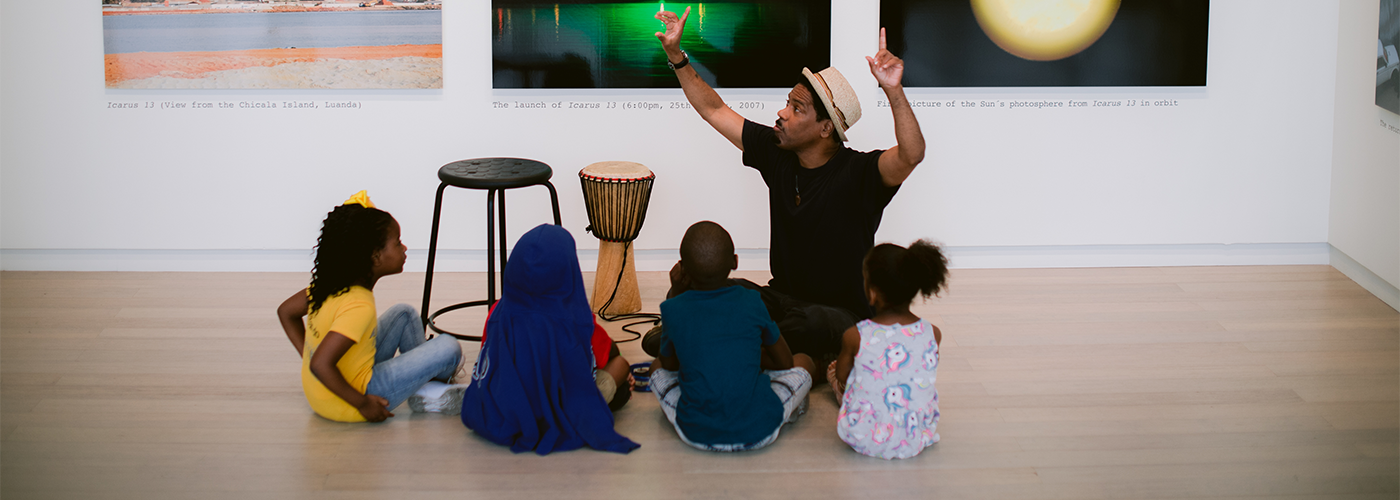 Four children seated around a gallery educator who is gesturing upwards. An African drum is to the educator's right.