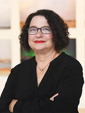 Bust portrait of Betti-Sue Hertz, Director and Chief Curator of the Wallach Art Gallery. The Director has chin length dark curly hair and is posed with her arms crossed. She is wearing glasses, a black v-neck top, and a short gold pendant with small golden sphere that falls just below her larynx.