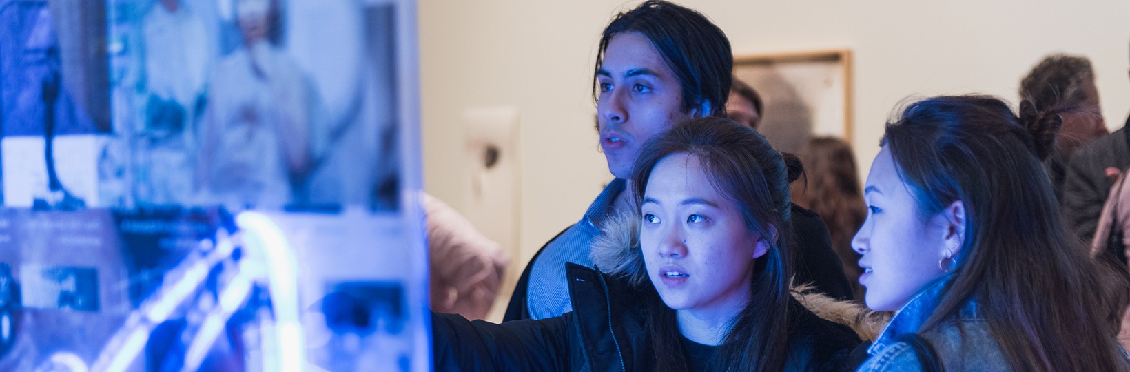 Three young individuals look at an artwork in a crowded exhibition space. 