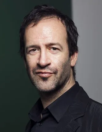 Portrait of Todd Shepard, head and shoulders. Dark hair, oval face with dark eyes and very short facial hair; wearing a black collared shirt and black blazer with a notched collar
