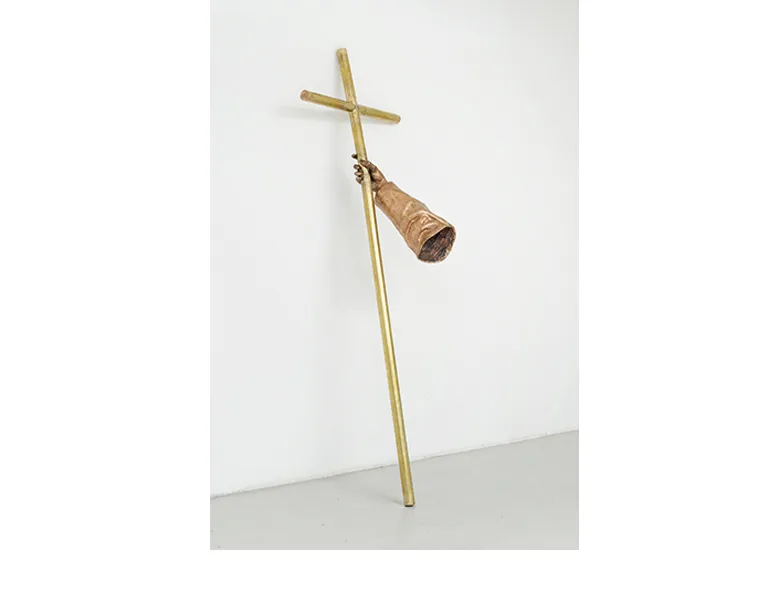 Sculpture by Fayçal Baghriche titled "Le bras du Cardinal (The Cardinal's Arm)." A bronze for arm holds a brass cross which leans against a white wall.
